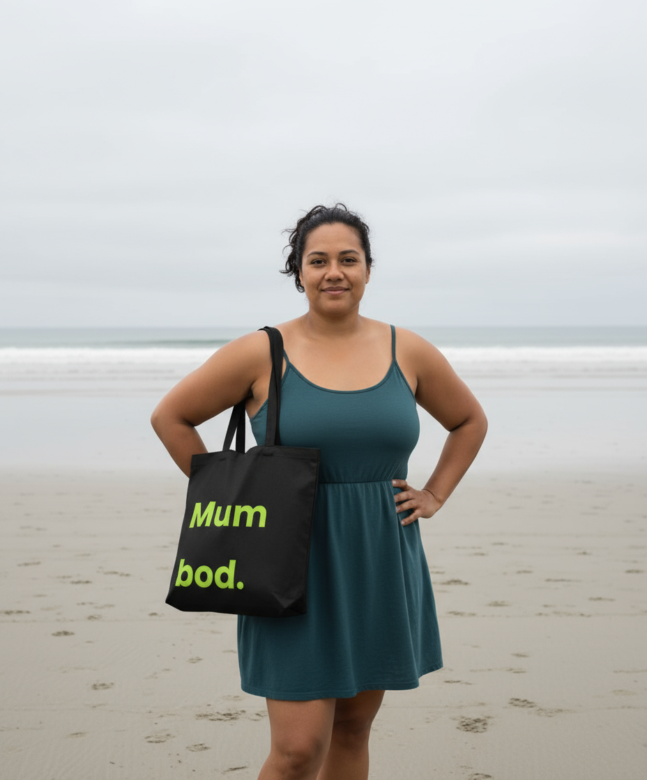 Woman holding a black tote bag with 'Mum bod.' text on a beach