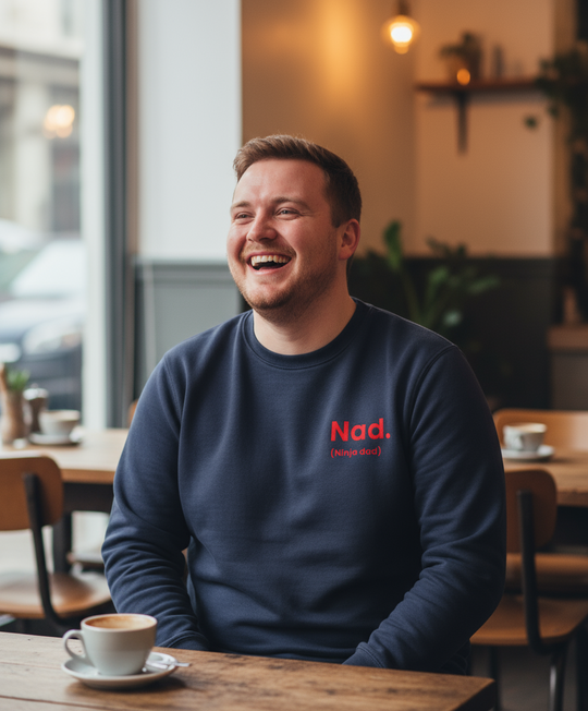 Man in a navy blue sweatshirt with 'Nad.' logo sitting at a table in a cafe, laughing.