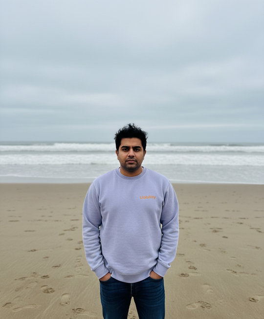 Man standing on a beach with light purple sweater and jeans, looking at the camera