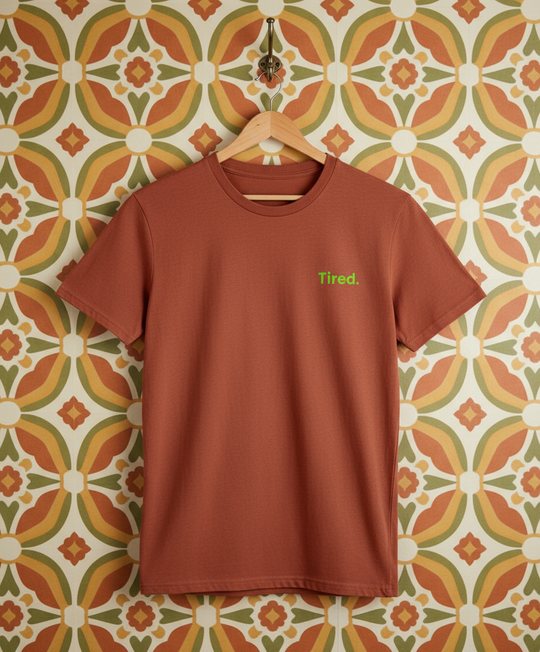 Brown t-shirt with 'Tired' text on a hanger against a patterned wall.