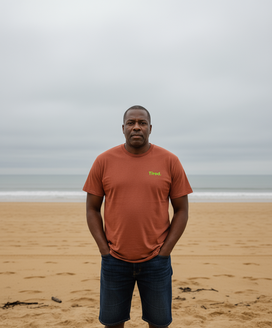 Man standing on a beach wearing an brown t-shirt with a logo.