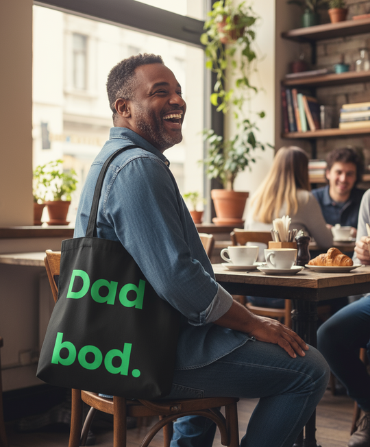 Man sitting in a cafe with a 'Dad bod.' bag, smiling.