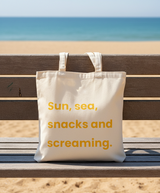 Beige tote bag with 'Sun, sea, snacks and screaming.' text on a beach bench with ocean view