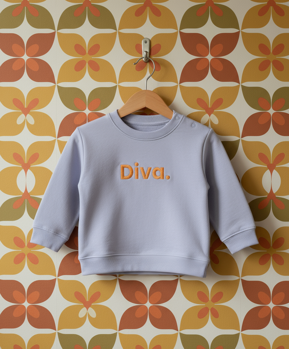 Light purple baby sweatshirt with 'Diva.' text on a hanger against a patterned wall.