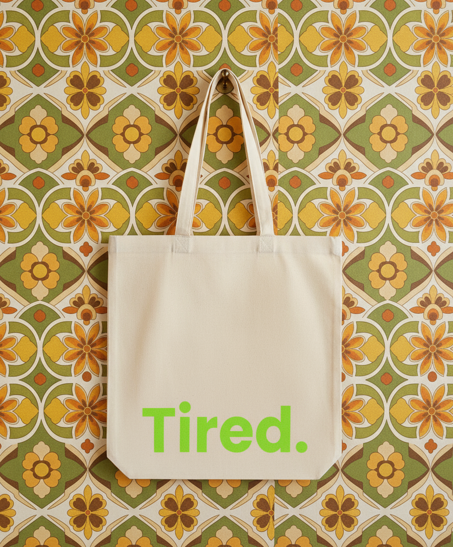 Beige tote bag with 'Tired.' text on a floral patterned wall.