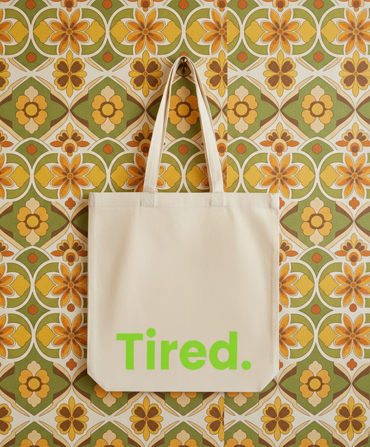 Beige tote bag with 'Tired.' text on a floral patterned wall.