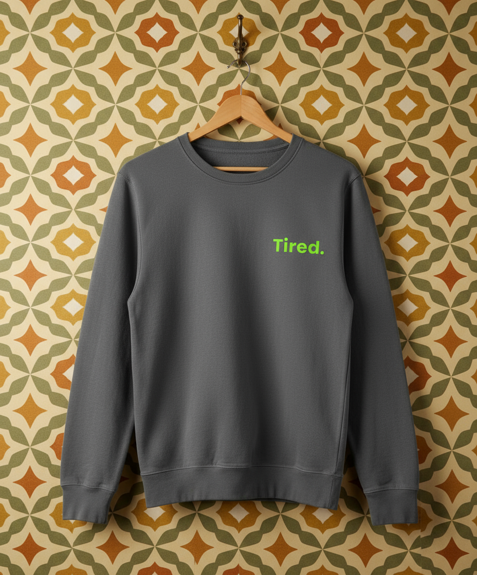 Grey sweatshirt with 'Tired.' text on a hanger against a patterned wall.