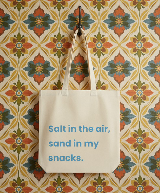 Tote bag with text 'Salt in the air, sand in my snacks' on a floral patterned wall.