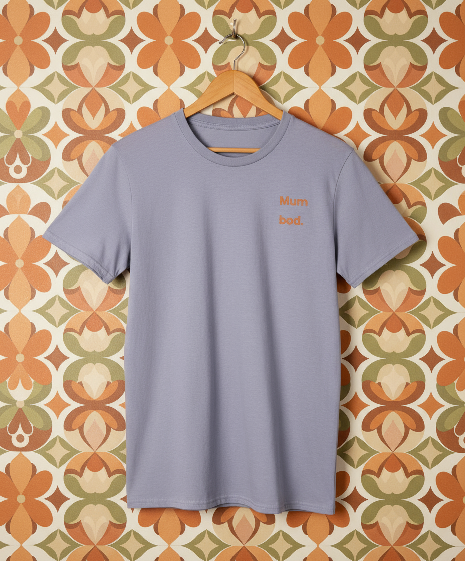 Light purple t-shirt with 'Mum bod' text on a hanger against a patterned wall.