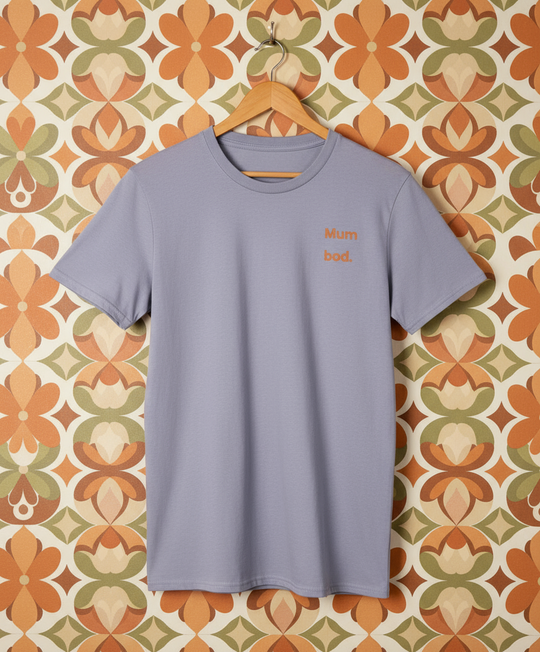 Light purple t-shirt with 'Mum bod' text on a hanger against a patterned wall.