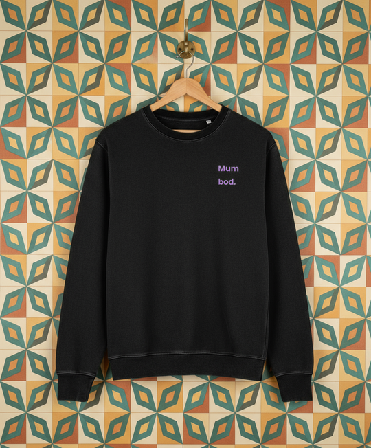 Black sweatshirt with 'mum bod' test hanging on a hook on a wall with retro wallpaper