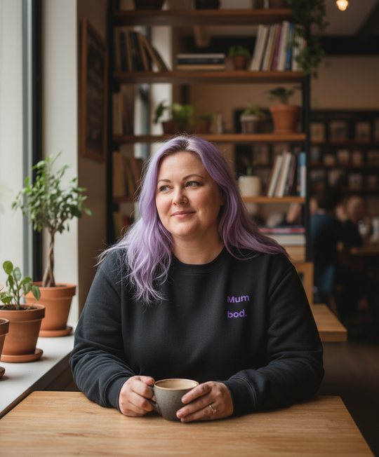 Woman sitting in cafe wearing black sweatshirt with 'mum bod' written on