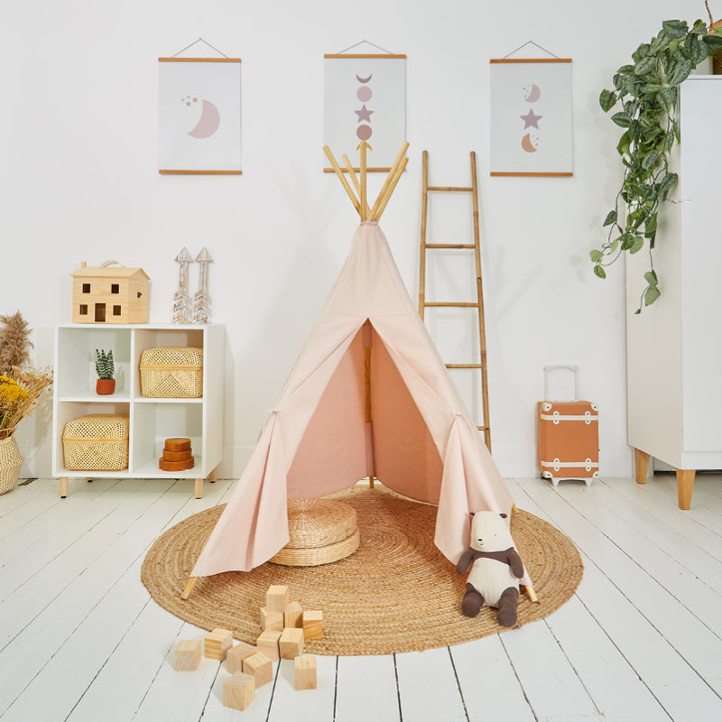 Children's play teepee in a room set up for play with wooden blocks, a doll, and a plant in the background. The teepee is pink and made from cotton canvas.
