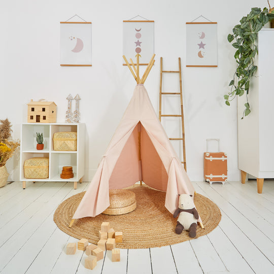 Children's play teepee in a room set up for play with wooden blocks, a doll, and a plant in the background. The teepee is pink and made from cotton canvas.