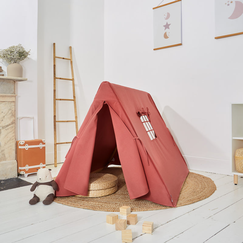 A pink play tent made from cotton canvas set up on a white room with a small wooden table and toys inside. There is a nine-paned window on the tent.