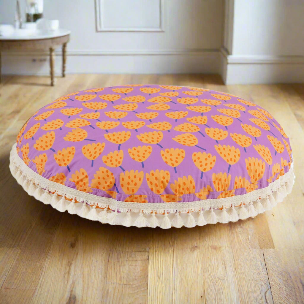 Round floor cushion with a purple background and an orange floral tulip pattern and white trim with tassels