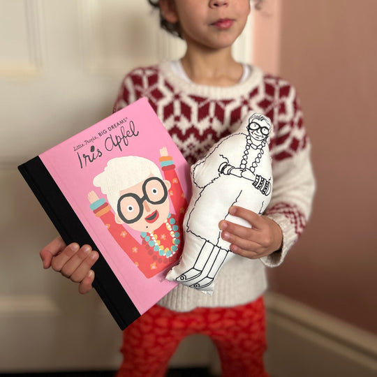 A child holding screen printed doll cushion and Little People Big Dreams book about Iris Apfel's life