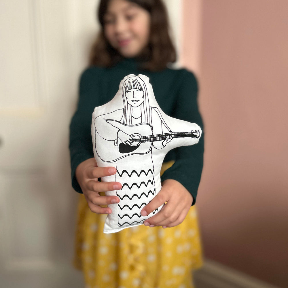 A child holding a black and white screen printed cushion doll depicting singer-songwriter Joni Mitchell playing a guitar.