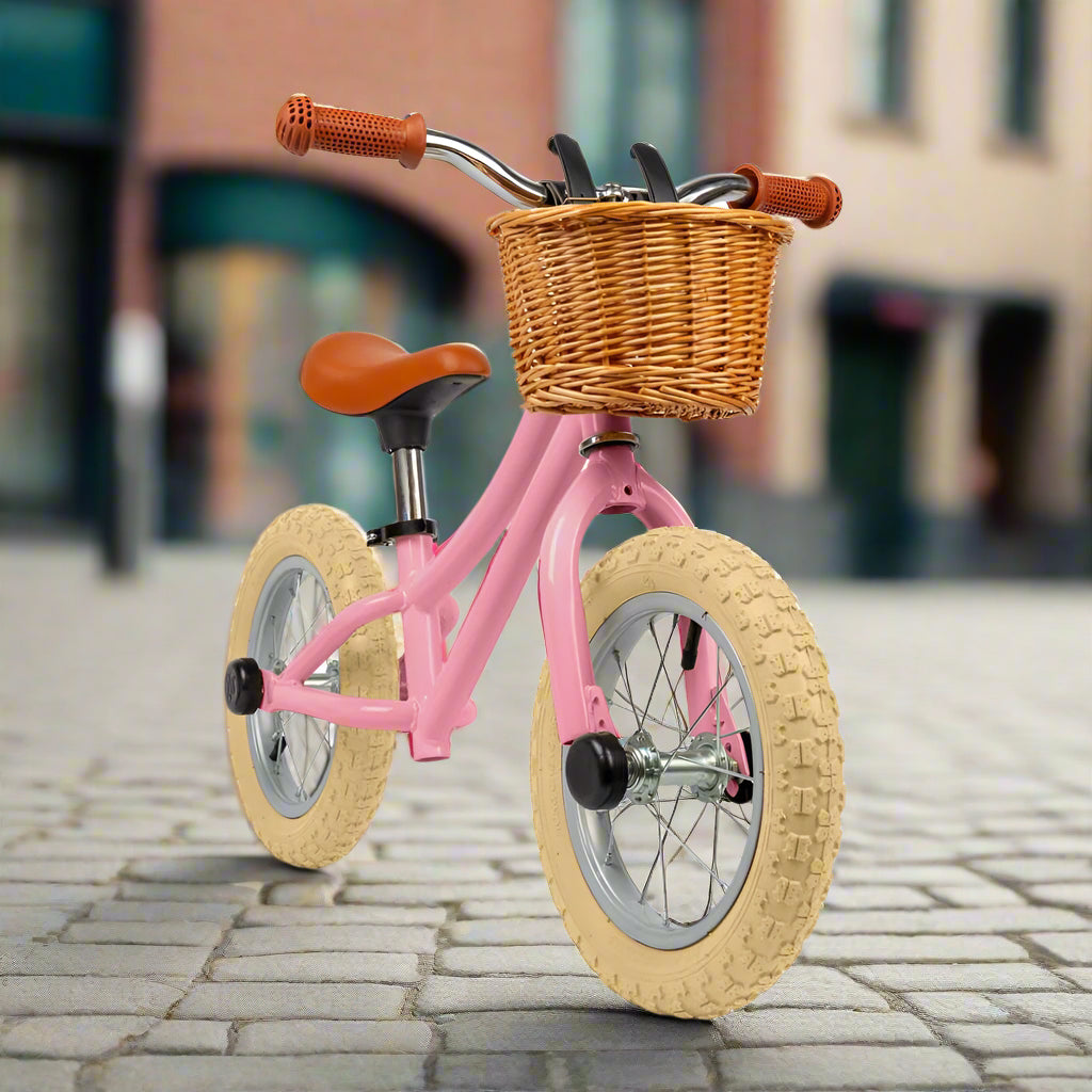 Balance bike with pale pink metal work and cream wheels with a brown saddle and brown handles with a brown wicker basket attached to the handlebar