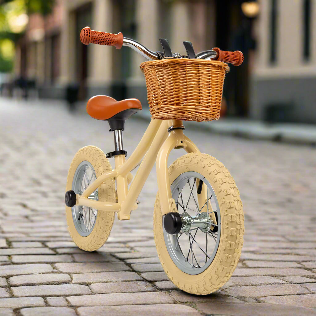 Cream coloured child's balance bike with cream wheels, brown seat, brown handles and a brown wicker basket attached to handlebars