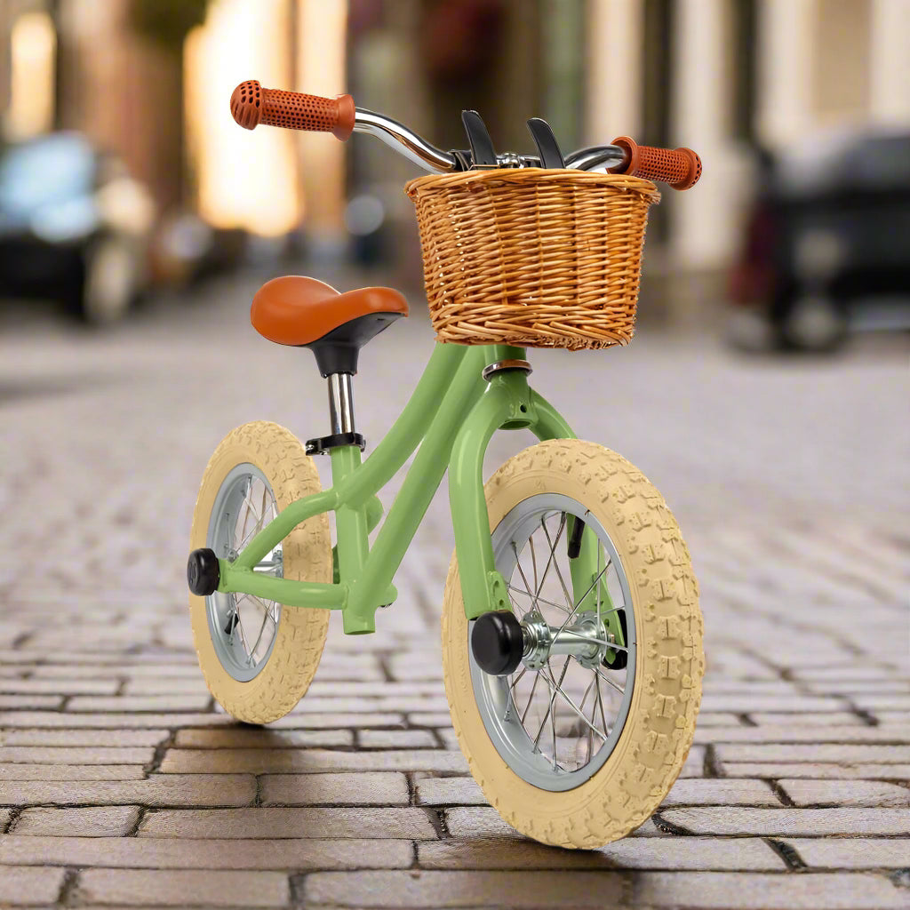 Balance bike with sage green metal work and cream wheels with a brown saddle and brown handles with a brown wicker basket attached to the handlebar