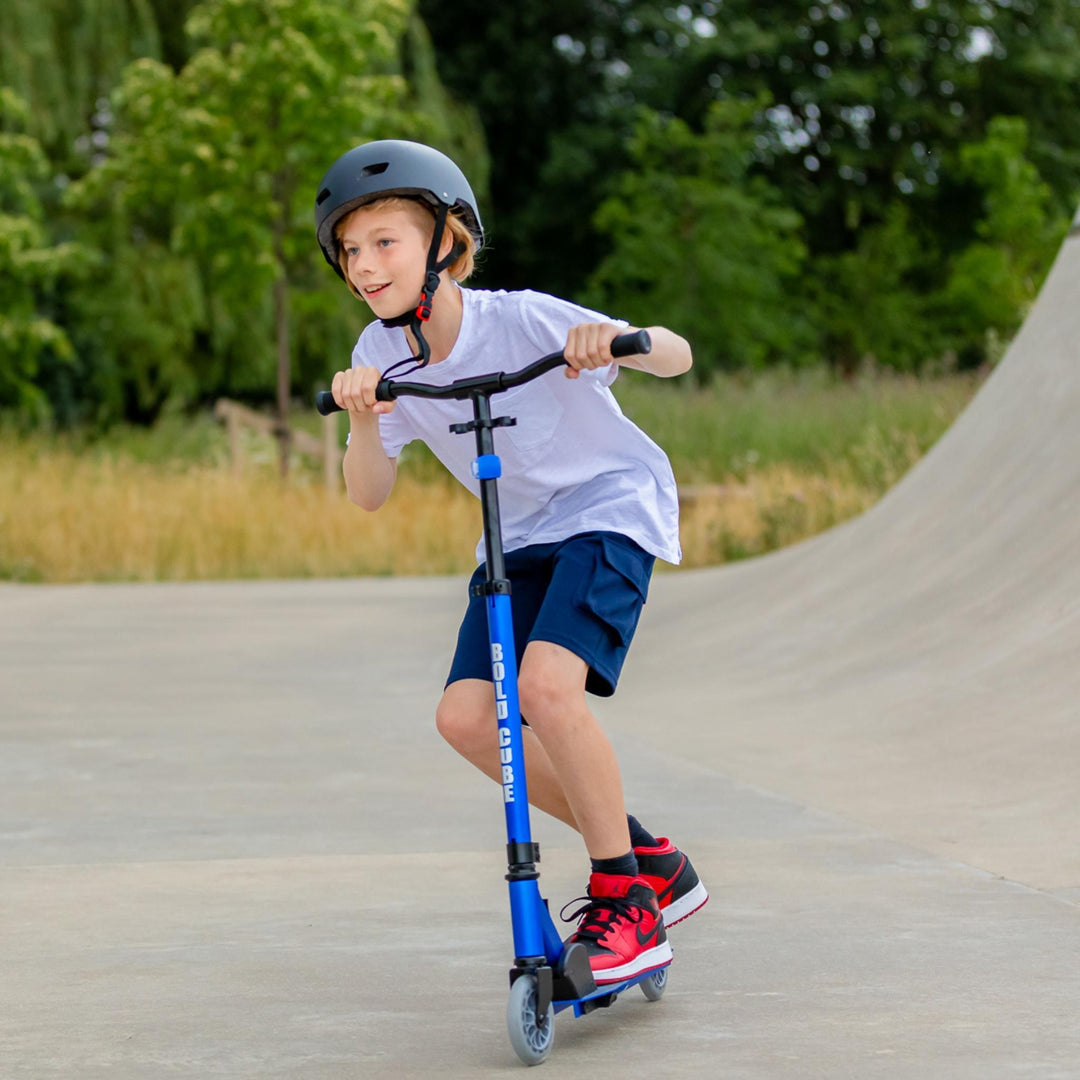 Child wearing a helmet riding a scooter in a skate park with greenery in the background