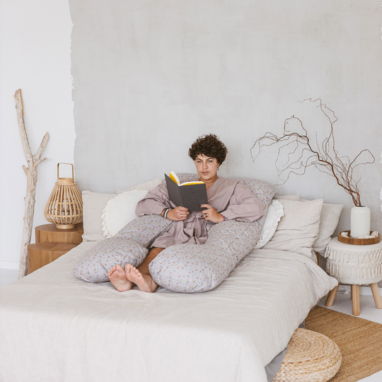 A person relaxing on a bed reading a book with the support pillow wrapped around their back