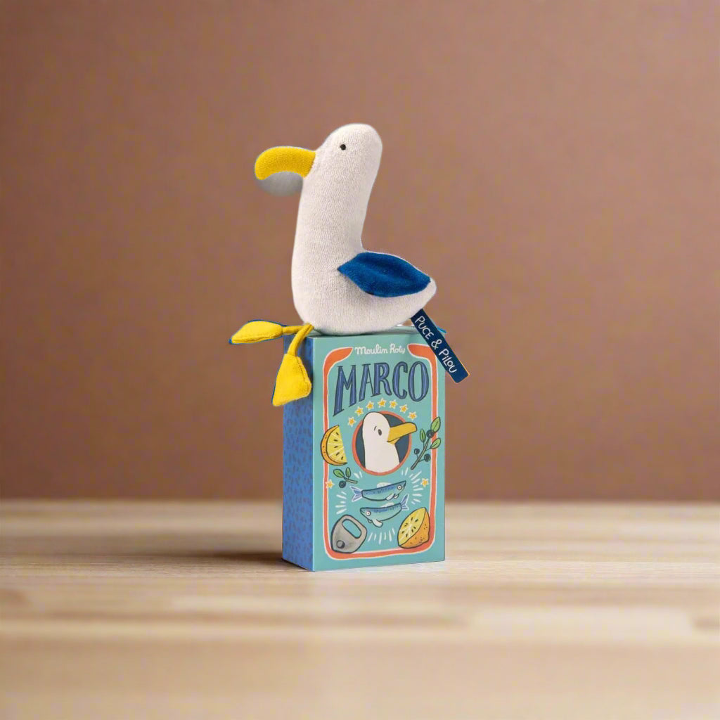 Plush seagull toy rattle with white body, yellow beak and feet and blue wings sitting on its decorative packaging box