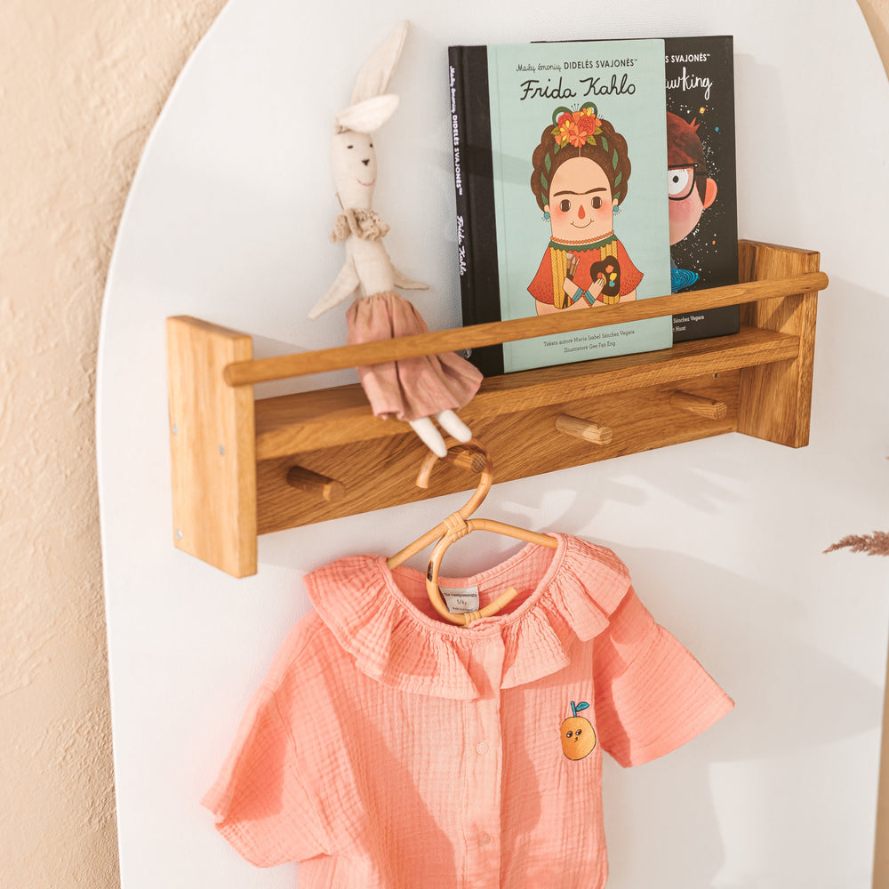 Side angle of the wooden shelf with a toy and two books on it, with wooden pegs underneath and a pink top hanging on the peg
