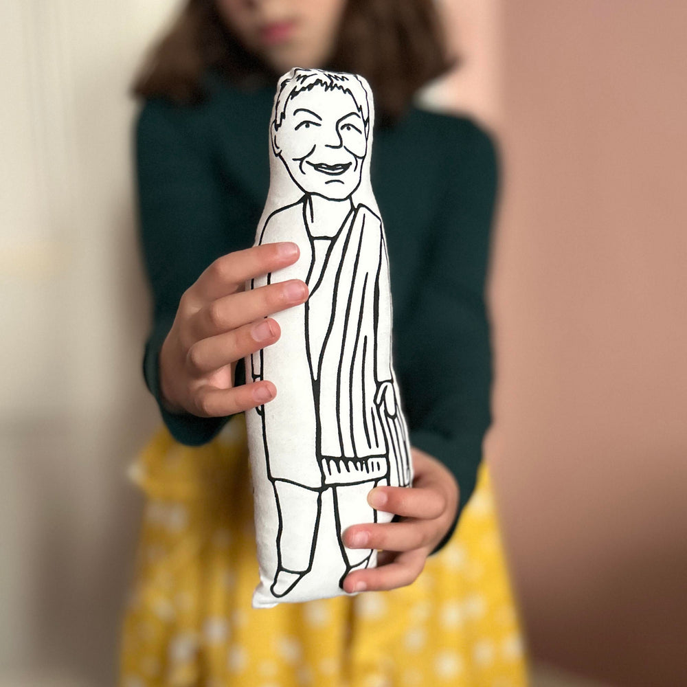 A child holding out a screen printed cushion doll depicting Dame Judi Dench towards the camera with an out of focus background
