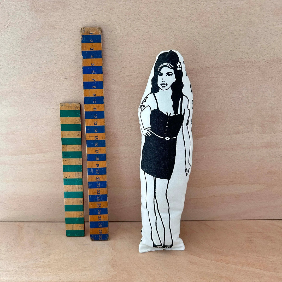 Screen printed doll cushion with white fabric and black illustration depicting singer songwriter Amy Winehouse wearing a black strap dress with one hand on her hip