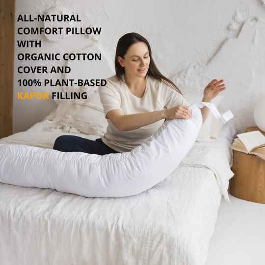 Woman sitting on a bed with an all-natural comfort pillow featuring organic cotton cover and plant-based kapok filling.