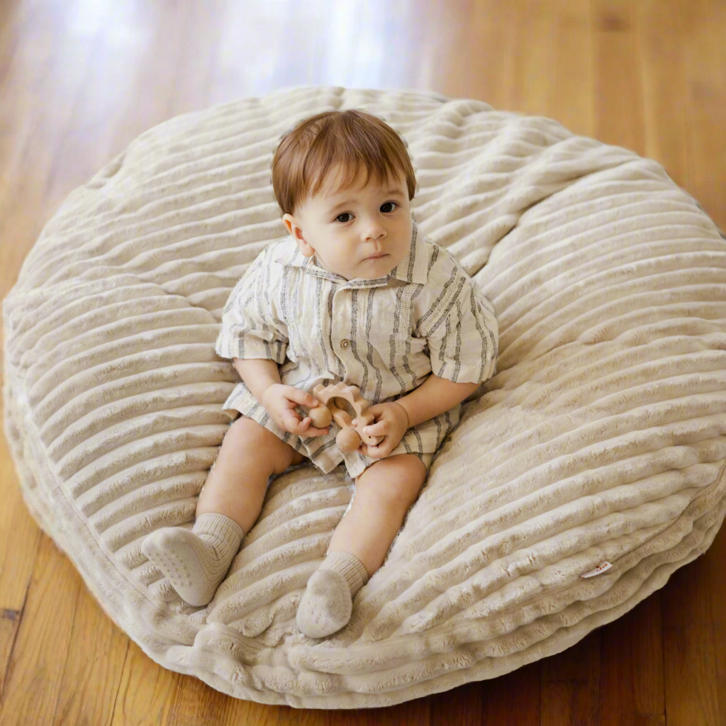 Young child sitting on a large beige textured floor cushion holding a toy