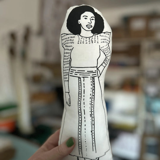 A hand holding up a screen printed doll cushion of Chimamanda Ngozi Adichie with a blurred background