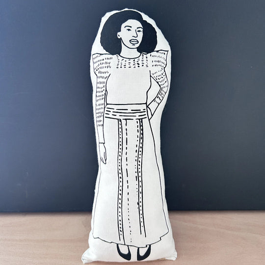 Screen printed doll cushion with white fabric and black illustration depicting author, feminist and TED speaker Chimamanda Ngozi Adichie wearing a long traditional dress with one hand on her hip