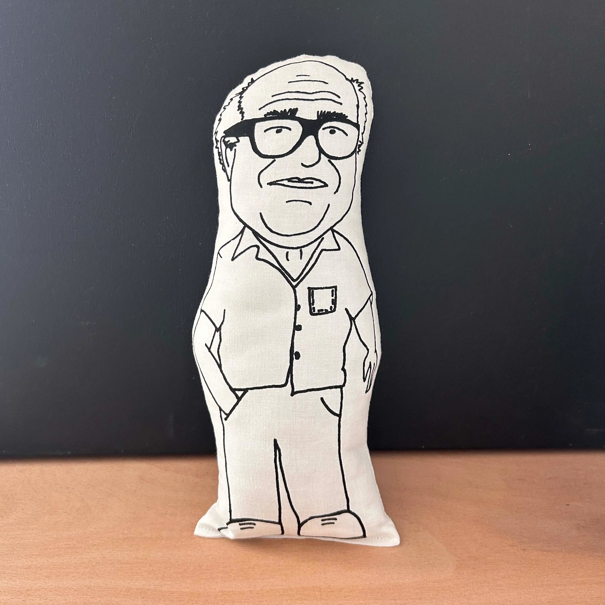 Screen printed doll cushion with white fabric and black illustration depicting actor and director Danny DeVito, wearing a short sleeved shirt, with one hand in his pocket and wearing thick rimmed glasses