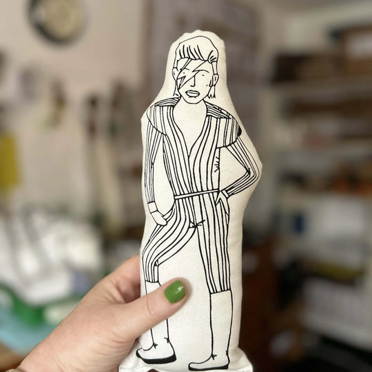 Hand holding a screen printed doll cushion with black line art of David Bowie against a blurred indoor background
