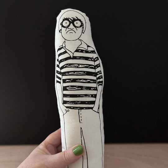 A hand holding the screen printed doll cushion of David Hockney against a dark background