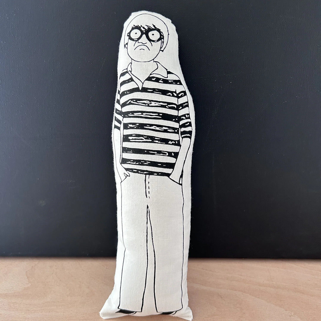 Screen printed doll cushion with white fabric and black illustration depicting artist David Hockney wearing loose trousers and a striped rugby-style top, with large round glasses and his hands in his pockets