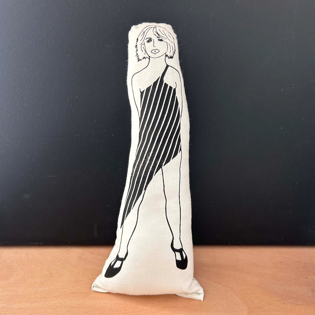 Screen printed doll cushion with white fabric and black illustration depicting musician Debbie Harry wearing a one-shoulder a-symetrical dress with diagonal stripes and heeled shoes