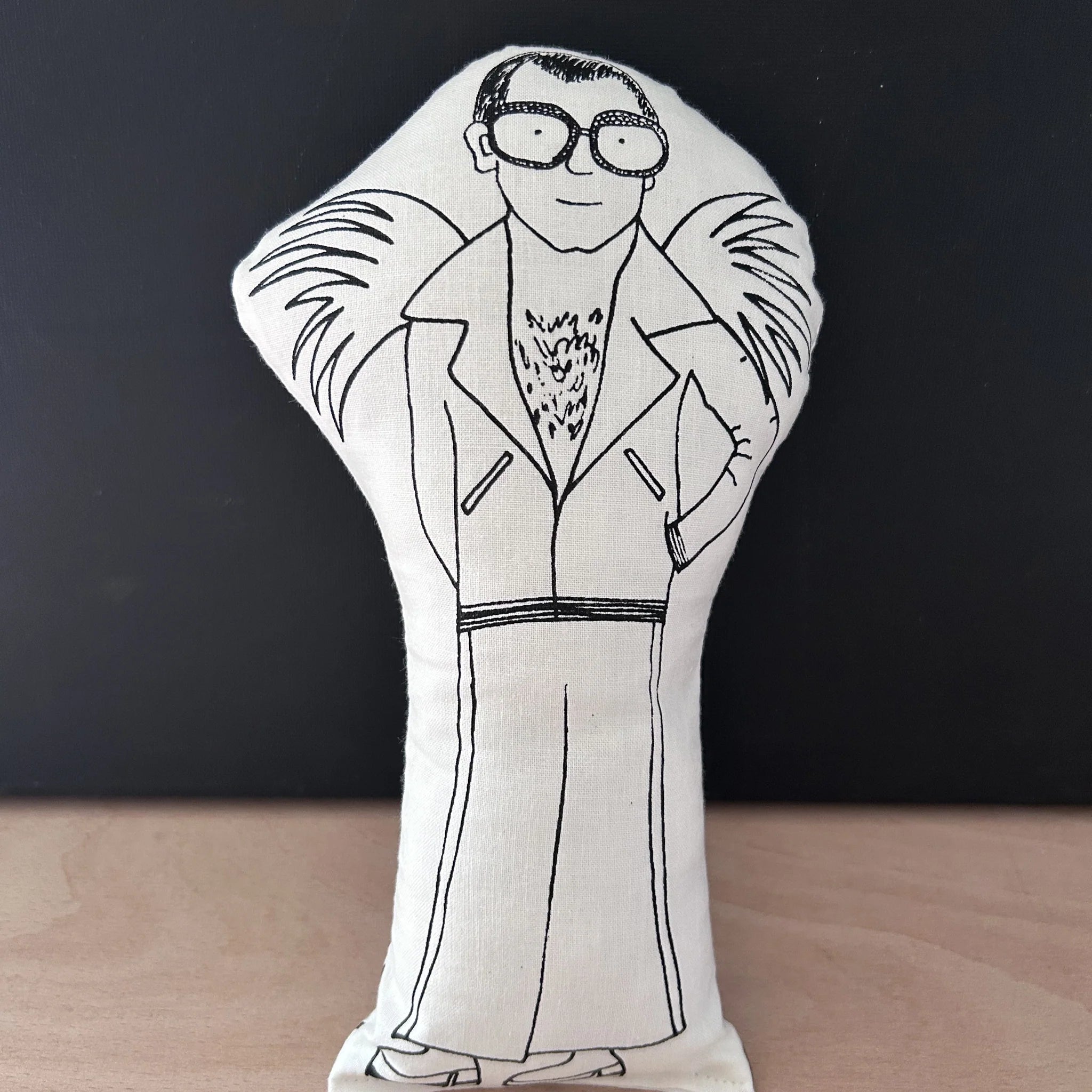 Decorative doll/cusion in white fabric with a black drawn illustration of a Elton John wearing large framed glasses and a suit with large lapels and heeled shoes
