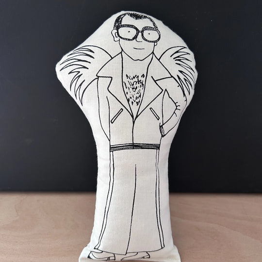 Decorative doll/cusion in white fabric with a black drawn illustration of a Elton John wearing large framed glasses and a suit with large lapels and heeled shoes