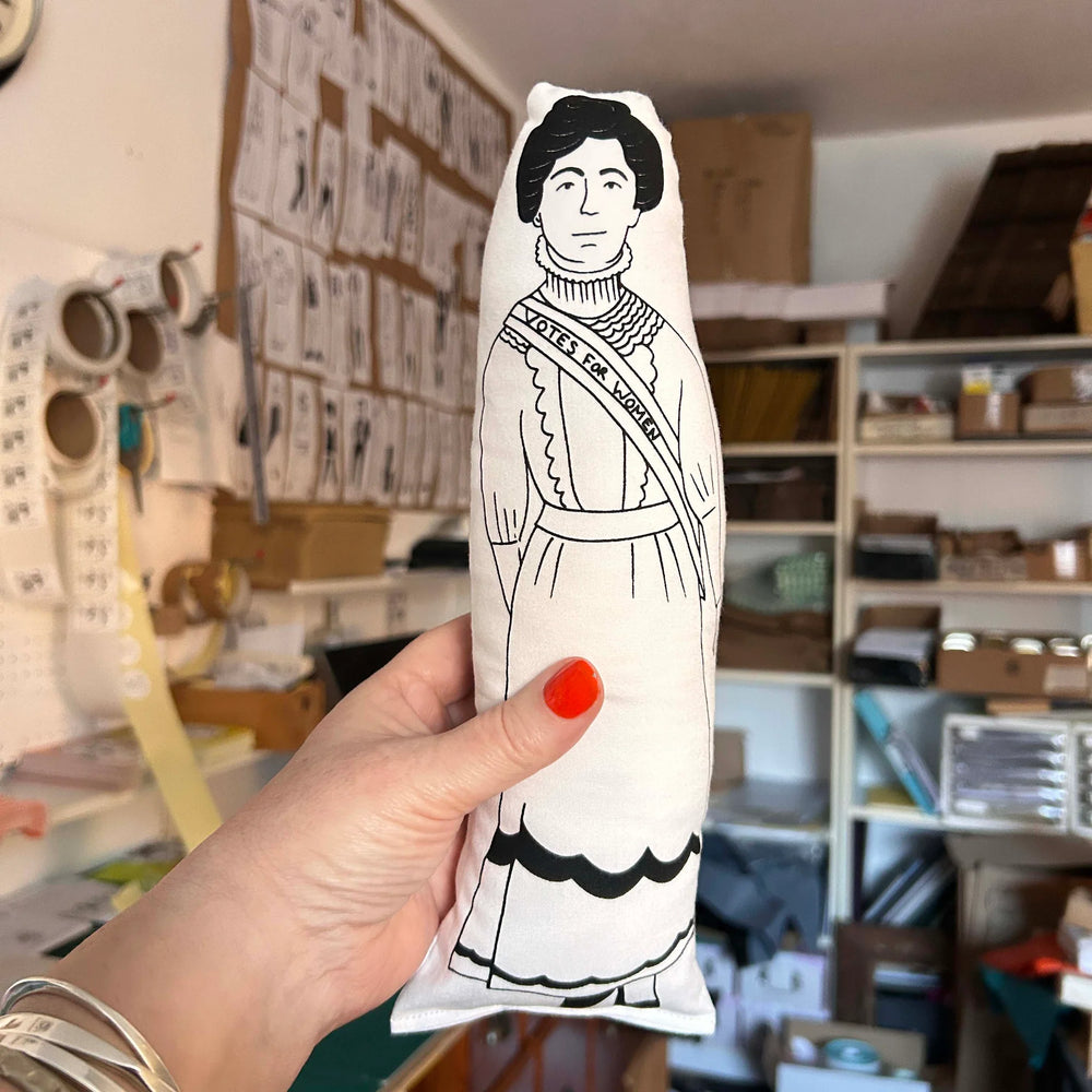 A hand holding a screen printed doll cushion of activist Emmeline Pankhurt with an out of focus backdrop of an interior room
