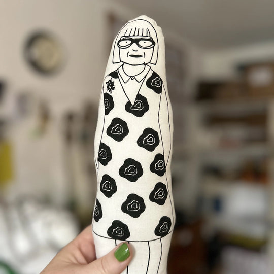 A hand holding up the screen printed doll cushion of The Sewing Bee judge Esme Young with a blurred background