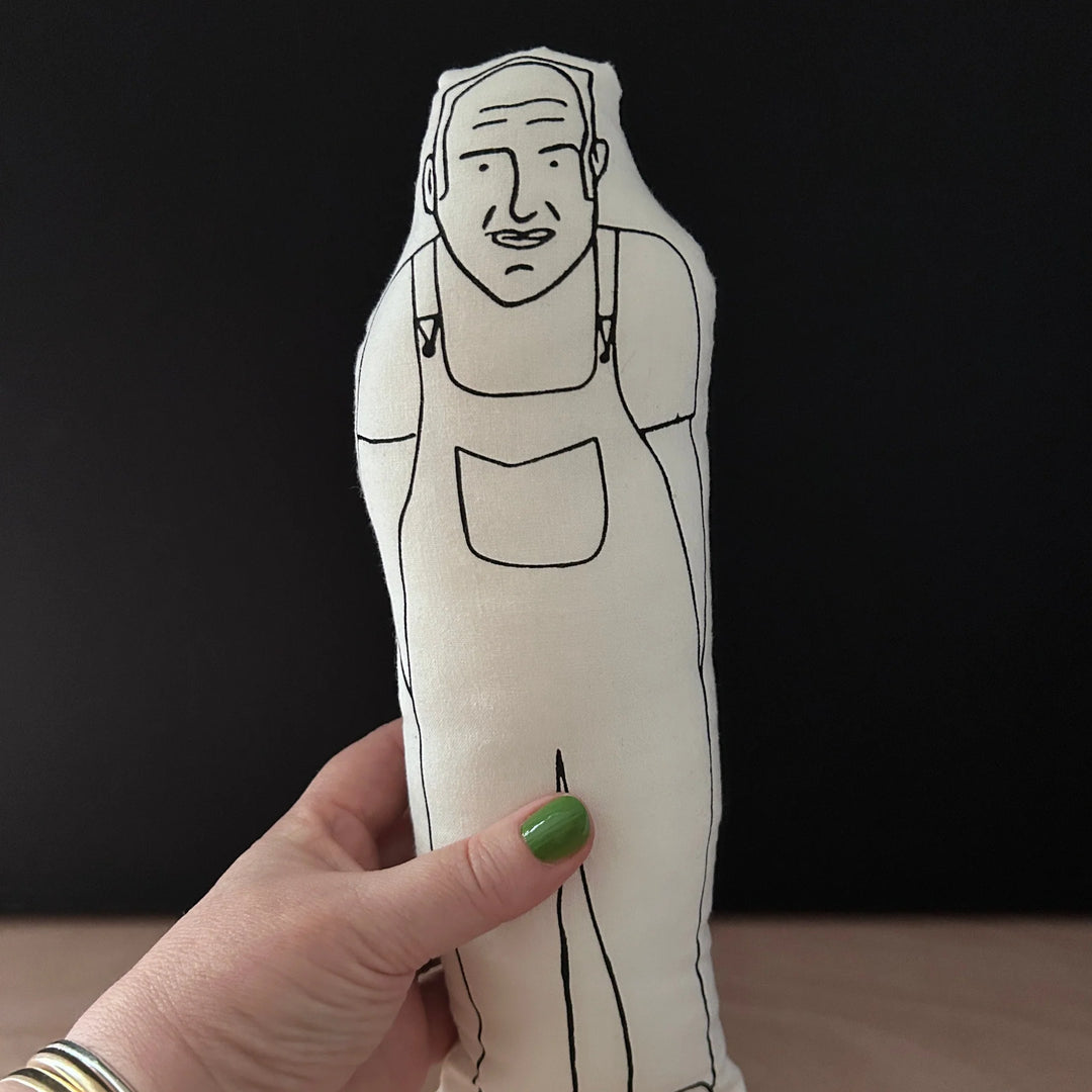 Hand holding a screen printed doll cushion with white fabric and black illustration depicting judge of The Great Pottery Throw Down - Keith Brymer Jones