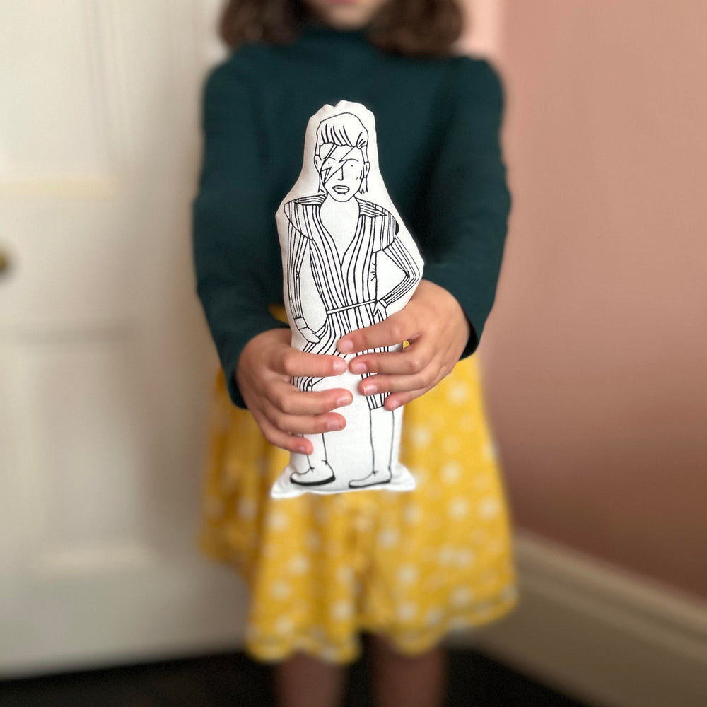 A child holding out the screen printed doll cushion of David Bowie as Ziggy Stardust towards the camera with a blurred background