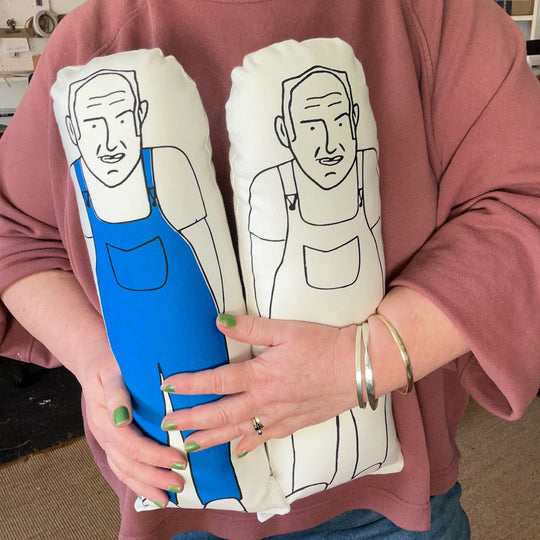 A person holding two large 45cm sized screen printed doll cushions of Keith Brymer Jones, one in black and white and one coloured with the overalls coloured blue