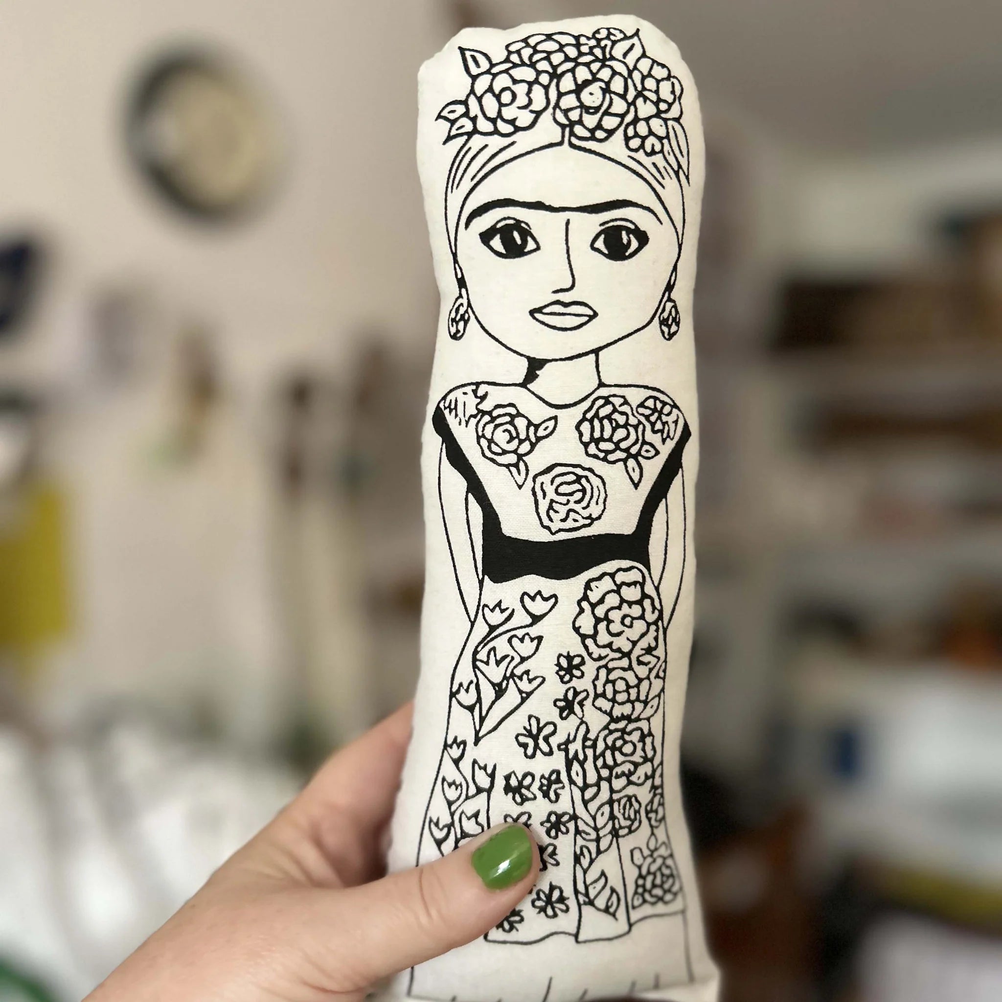 Hand holding a screen printed doll cushion with white fabric and black illustrations depicting he artist Frida Kahlo wearing a floral traditional dress