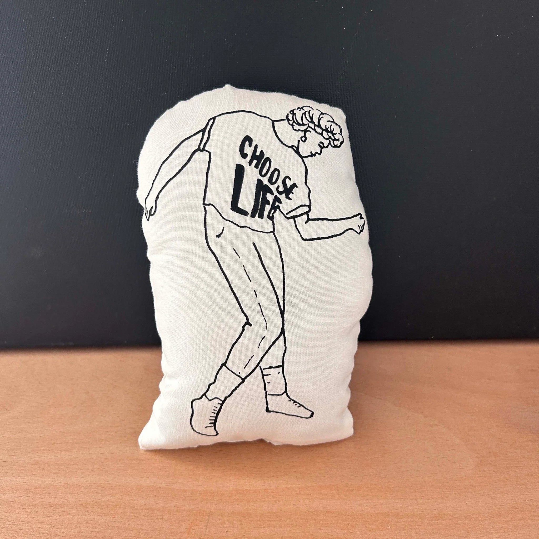 Screen printed doll cushion with white fabric and black illustration depicting singer-songwriter George Michael dancing wearing a 'Choose Life' T-shirt