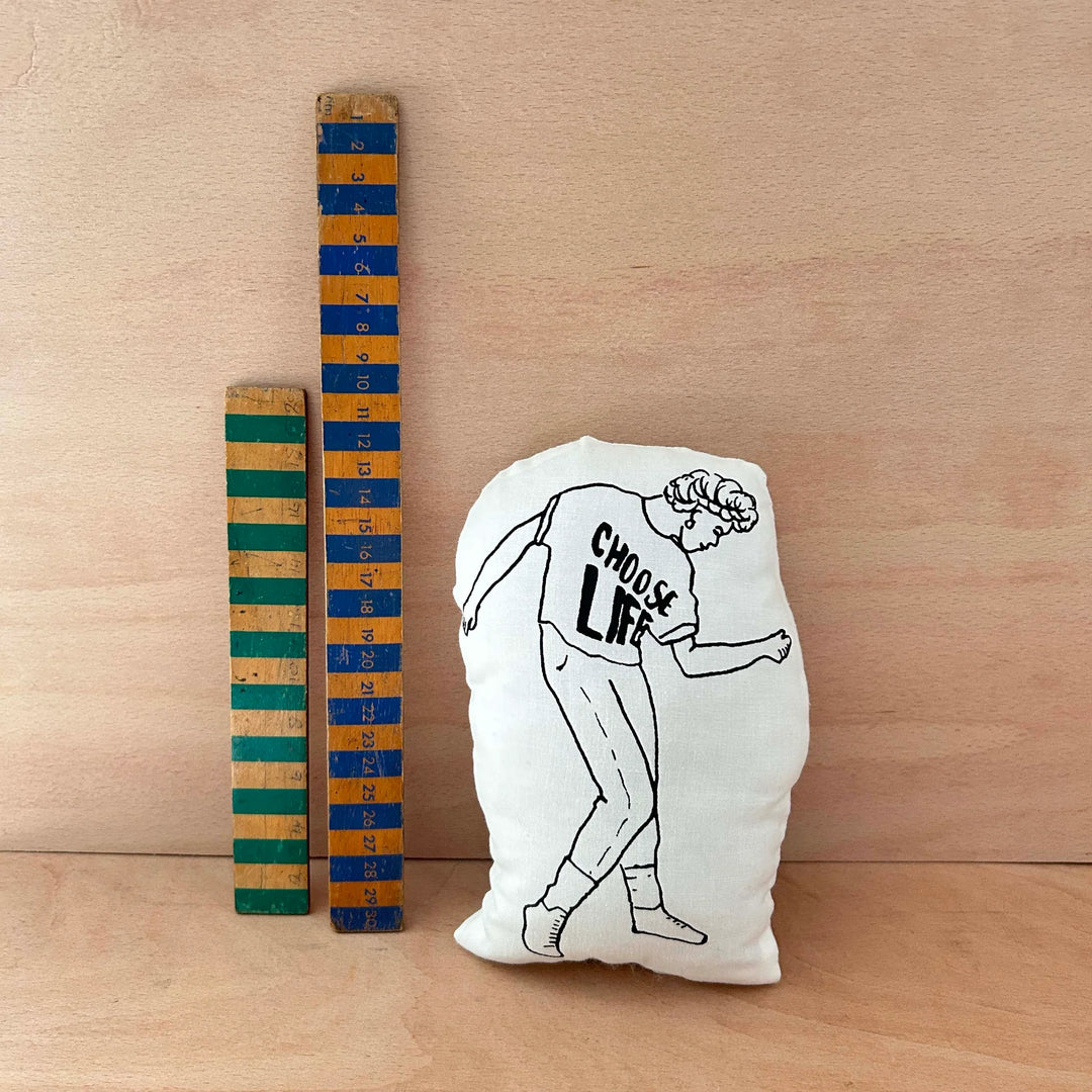Screen printed doll cushion of George Michael dancing next to two wooden rulers on a wooden background
